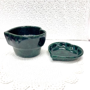May include: A set of two heart-shaped ceramic dishes in a deep green color. The larger dish is taller and has a wider opening, while the smaller dish is shallower and more compact.