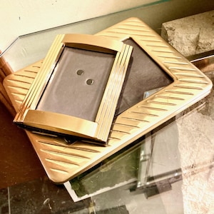 May include: Two rectangular gold-toned metal picture frames with a diagonal ribbed texture on the borders. The smaller frame is positioned at an angle on top of the larger frame, both featuring dark gray backing inserts and smooth metallic edges.