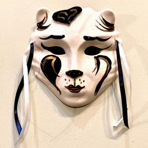 May include: A white ceramic mask in the shape of a cat's face, adorned with black and gold accents. The mask features closed eyes, a small nose, and a detailed mouth. Ribbons in white and navy blue hang from the sides.