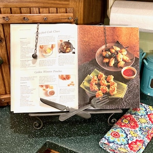 May include: An open cookbook on a decorative metal stand, displaying recipes for crab claws and wonton pouches. A fork and knife rest on the stand. The cookbook is held open by a chain. A colorful floral napkin is in the foreground.