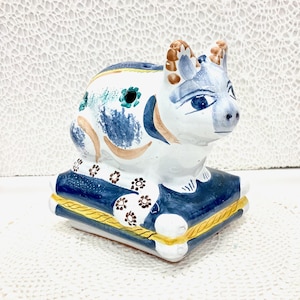 May include: A ceramic figurine of a stylized animal, possibly a cow or bull, in white, blue, and brown. The figurine is seated on a blue cushion with gold trim. Decorative floral and abstract patterns adorn the animal's body.