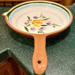 May include: A ceramic serving dish with a handle. The dish has a brown rim and handle, with a floral design in the center featuring yellow, purple, and green flowers. The rim is decorated with blue and green accents.