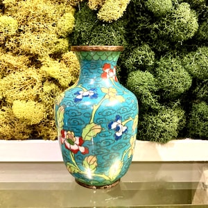 May include: A turquoise cloisonné vase with floral designs in red, white, and blue. The vase has a gold rim and base, and is set against a backdrop of yellow and green moss.