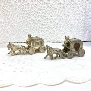 May include: Two silver-colored miniature stagecoaches, each pulled by a team of two horses. The stagecoaches have detailed wheels and a driver figure. The background is a white, textured surface.