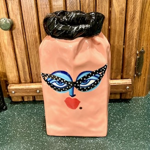 May include: A pink ceramic decorative jar with a face design. The face features blue eyes, black and white polka dot glasses, red lips, and a beauty mark. The top of the jar is black.