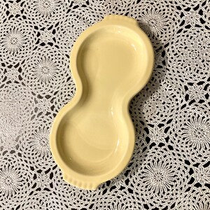 May include: A pale yellow ceramic serving dish shaped like a peanut with two rounded sections and a smooth, glossy finish. The dish features small, ridged handle-like extensions at both the top and bottom ends, creating a symmetrical form.