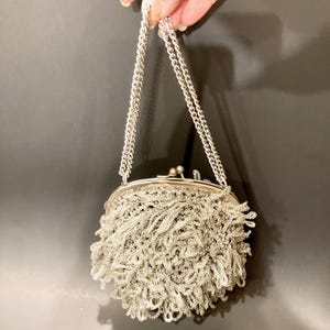 May include: A silver-colored, vintage-style purse with a chain strap. The purse has a clasp closure and is adorned with a fringe of silver threads, creating a textured, decorative effect. The chain strap is also silver.