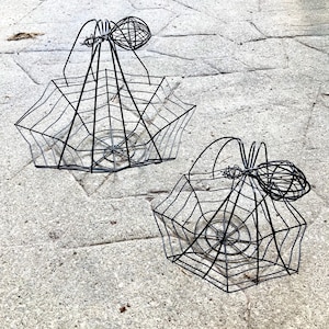 May include: Two black wire baskets with a spiderweb design. Each basket has a handle and a spider-shaped accent. The baskets are made of thin metal rods and are designed to resemble a spiderweb.