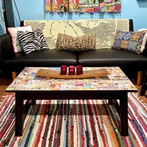 May include: A dark brown coffee table with a collage top, holding three red votive candle holders and a wooden tray. The table sits on a colorful striped rug, with a black sofa and decorative pillows in the background.