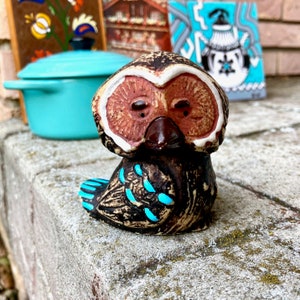 May include: A ceramic owl figurine with brown and white paint, blue accents, and a brown beak. The owl is sitting on a gray surface.