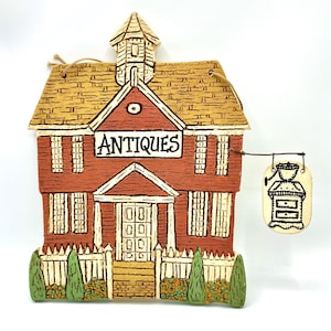 May include: A ceramic antique shop ornament. The building is red with a tan roof and white trim. The word "ANTIQUES" is displayed on a sign above the door. A small sign with a dresser hangs from the side.