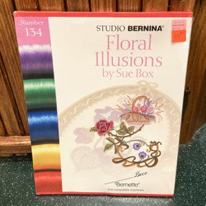 May include: A sealed package for Studio Bernina Floral Illusions by Sue Box. The package features a floral design with a basket of flowers and the word "Deco". The package also has a number 134 on the side and a variety of colored threads.