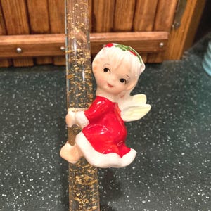 May include: A vintage Christmas angel figurine in a red dress with white trim and small yellow wings. The angel is holding onto a clear glass tube filled with gold flakes. The angel has white hair and a green and red holly wreath.