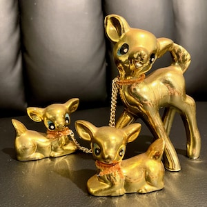 May include: A set of three gold-toned ceramic deer figurines with a shiny metallic finish. One larger standing deer is connected by thin gold chains to two smaller, reclining deer. Each deer features painted black and white eyes and a small orange collar.