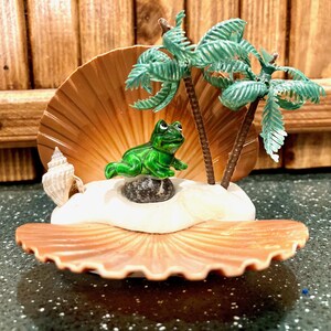 May include: A decorative seashell scene featuring a green frog on a small rock, miniature palm trees, and a small seashell. The frog is positioned on a white island within a large, open seashell.