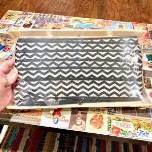 May include: A rectangular fabric pouch featuring a repeating black and white chevron pattern. The pouch is encased in a clear plastic protective sleeve and displays a consistent zigzag design across the entire front surface of the item.