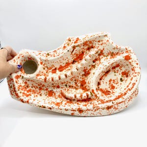 May include: A white ceramic tray with an irregular, organic shape featuring a vibrant orange and green paint splatter pattern. The tray includes a circular opening, a ridged section, and a recessed basin, all finished with a glossy glaze.