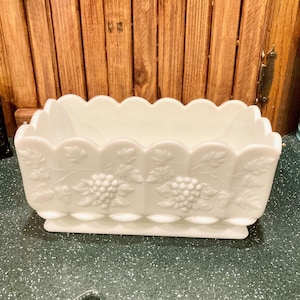May include: A rectangular, white, vintage glass dish with a scalloped rim. The dish features embossed grape and leaf designs on the sides. The base is rectangular and slightly raised, adding to its classic appeal. A decorative piece for serving or display.