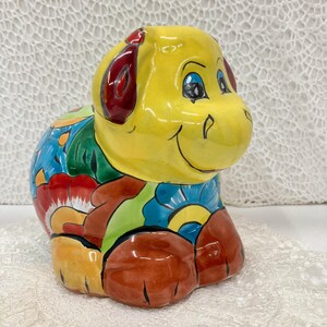 May include: A ceramic dog figurine with a yellow head and red ears. The body is decorated with colorful floral patterns in blue, red, green, and orange. The dog is sitting with its legs tucked under.