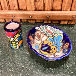 May include: A set of two colorful ceramic dishes with a blue rim and floral designs. The smaller dish is a shot glass and the larger dish is a shallow bowl.