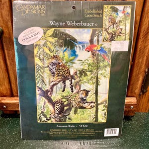 May include: A cross-stitch kit featuring a jungle scene with leopards, parrots, and a waterfall. The kit is titled "Amazon Rain" and includes the finished size of 12" x 16". The design is by Wayne Weberbauer.