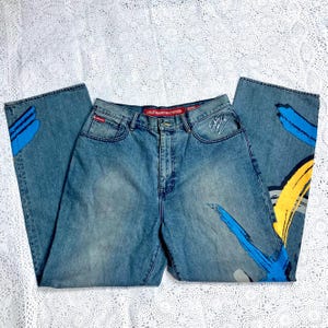 May include: Light blue denim jeans with colorful abstract paint strokes in blue, yellow, and gray. The jeans have a classic five-pocket design, a button and zipper closure, and the word "Entitled" is embroidered on the front pocket.