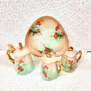 May include: A vintage porcelain condiment set featuring a triangular serving dish and three matching shakers. The set is painted with a floral design of red berries and green leaves on a pink and mint green background, with gold accents.