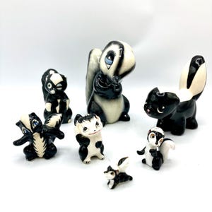 May include: A collection of seven ceramic skunk figurines in various poses, featuring glossy black and white painted details, expressive facial features with painted eyes, and raised tails, arranged together on a plain white surface.