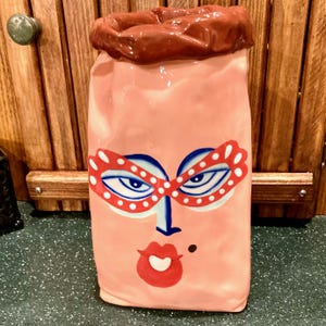 May include: A pink ceramic vase with a face design. The face features blue eyes, red-rimmed glasses, a red mouth, and a small black beauty mark. The top of the vase is brown.