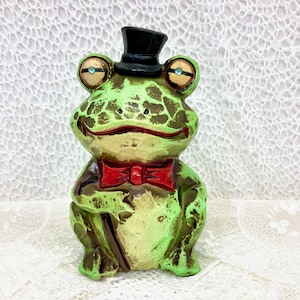 May include: A green frog figurine wearing a black top hat and a red bow tie. The frog is holding a brown walking stick and has a whimsical expression.
