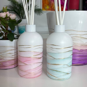 Op de afbeelding: Twee witte diffuserflessen met gouden accenten en roze en blauwe aquarelontwerpen. Elke fles heeft een set witte diffuserstokjes.