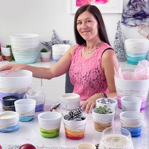May include: A woman sits at a table with a variety of white ceramic planters. The planters are decorated with gold and colourful abstract designs. Some of the planters have candles inside.