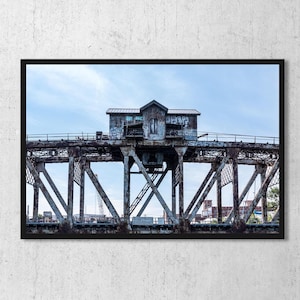 May include: A framed wall art print featuring a weathered, rusted metal bridge structure with a central control house covered in graffiti. The industrial bridge is set against a pale blue sky with light clouds, showcasing intricate steel trusses.
