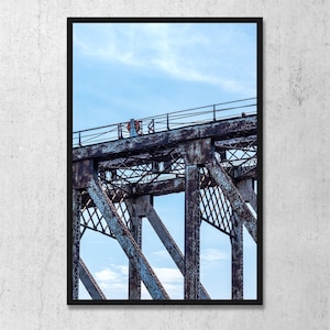 May include: A framed wall art print featuring a low-angle view of a weathered, rusted metal truss bridge structure against a light blue sky with wispy clouds. The bridge features dark brown and orange oxidation patterns and a small orange life ring.
