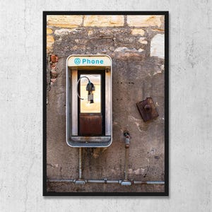 May include: A framed photograph of a vintage payphone mounted on a weathered concrete wall. The phone booth is silver with a blue "Phone" sign. The wall is a mix of concrete and stone, with metal fixtures and pipes.