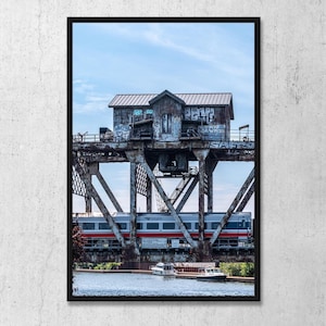 May include: A framed vertical photograph of a weathered, graffiti-covered metal lift bridge structure over a waterway. An Amtrak passenger train with red and blue stripes crosses the bridge, while two small white boats float on the water below.