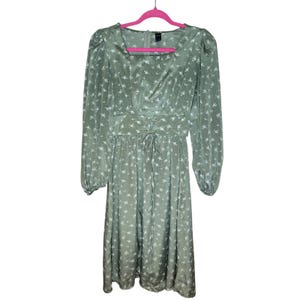 May include: A sage green midi dress with a white floral print and a square neckline. The dress has long sleeves with puffed shoulders and a fitted bodice with a lace-up detail at the waist.