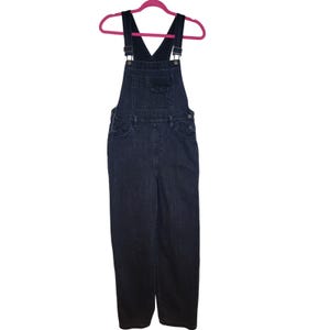May include: Dark wash denim overalls hanging on a pink hanger. The overalls have adjustable straps, a bib pocket, and side pockets. The denim is a deep indigo color, and the overall style is casual and relaxed.