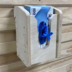 May include: A wooden wall-mounted glove dispenser. The dispenser is made of light-colored plywood and has a curved top. A box of blue gloves is partially visible inside, with a single blue glove extending out of the dispenser's opening. The box reads "100 Count by Weight".