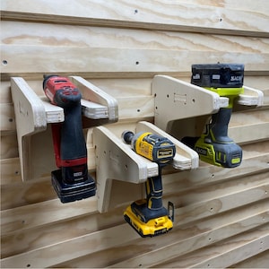May include: Three power drill holders mounted on a wood wall. The holders are made of wood and have a slot for the drill to fit into. The drills are a red and black drill, a yellow and black drill, and a green and black drill.