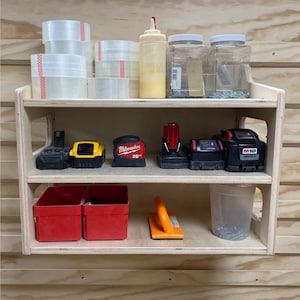 May include: A two-shelf wooden storage unit with various tools and supplies. The top shelf has clear jars with screws, a bottle of glue, and rolls of tape. The bottom shelf has two red plastic storage bins, a yellow tape measure, a clear plastic container with screws, and a yellow and black power tool battery.