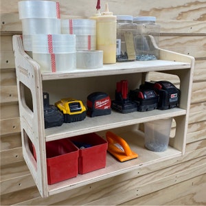 Puede incluir: Una estantería de pared de madera de dos niveles con varias herramientas y suministros. La estantería superior tiene rollos de cinta transparente, una botella de pegamento para madera y dos frascos de vidrio con tornillos. La estantería inferior tiene una batería de taladro amarilla y negra, una batería de taladro roja y negra, una batería de taladro roja y negra, una cinta métrica amarilla y negra, dos contenedores de plástico rojo y un contenedor de plástico transparente con tornillos. La estantería está hecha de madera clara y tiene un acabado natural.