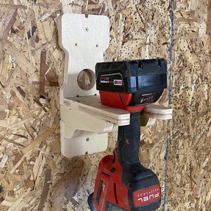 May include: A wooden wall mount for a cordless drill and battery. The mount is made of plywood and has a hole for the drill bit and a shelf for the battery. The drill is red and black and has the word "FUEL" on it. The battery is black and red and has the word "M18" on it.