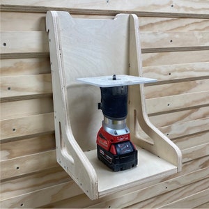 May include: A wooden wall mount for a cordless router. The mount has a shelf and a circular cutout for the router base. A red and black cordless router is mounted on the shelf.