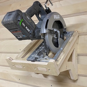 May include: A wooden wall mount for a circular saw with a black and green battery-powered saw attached. The saw has a silver blade with the word "FLEX" printed on it. The wall mount has a slot for the saw blade and a hook for the saw's handle.