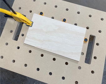 Op de afbeelding: Een stuk multiplex is met gele klemmen vastgezet op een werkbank met een prikbord. Het prikbord heeft een reeks gelijkmatig verdeelde gaten.