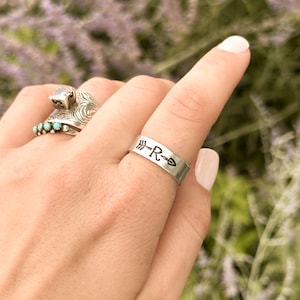 May include: A silver ring with an arrow, the letter "R", and a stylized fish symbol. The ring is on a finger, with other rings in the background. The background is a soft focus of purple flowers.
