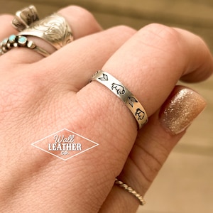 May include: A silver ring with a design of buffalo silhouettes and arrows. The ring is on a person's finger.