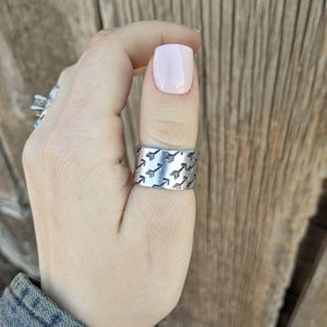 May include: A silver thumb ring with a repeating pattern of arrows. The ring is worn on a person's thumb.