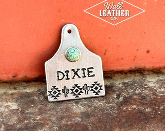 May include: A silver-toned metal livestock tag featuring a circular turquoise-colored stone at the top. The name DIXIE is stamped in black lettering above a row of black stamped geometric patterns and cactus icons. A small hole is at the top.